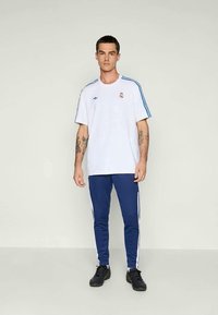 Tetovált férfi fehér Adidas Real Madrid pólót és kék Adidas melegítőnadrágot visel, míg egy sima világos falnak támaszkodik.
