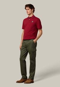 Rode polo met korte mouwen en kraag, donkere accenten en een logo op de borst; gecombineerd met groene cargobroek en bruine suède schoenen. Neutrale achtergrond.