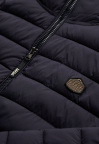 Gros plan sur une veste matelassée noire avec une fermeture éclair et un écusson brodé en forme d'hexagone portant le logo "Superdry Outdoor".