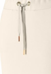 Der hellbeige Sweatshirtstoff verfügt über einen elastischen Bund mit Kordelzug, der mit goldakzentuierten Metallspitzen an den Bändern verziert ist.