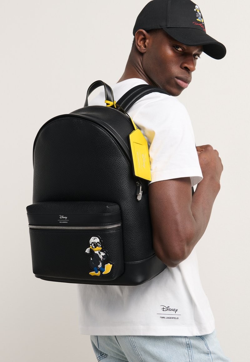 Melnā teksturētā ādas mugursoma ar priekšējo rāvējslēdzēja kabatu, kurai ir dzeltena birka un karikatūras Džona Duka grafika priekšpusē.