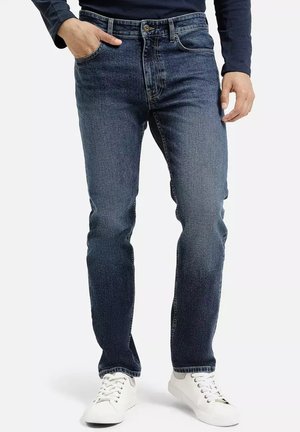 Ciemnoniebieskie dżinsy o slim fit, z pięcioma kieszeniami, zapięciem na guzik i widocznym szwami. Noszone z białymi trampkami.
