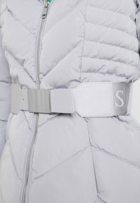 Veste matelassée gris clair avec fermeture éclair argentée et large ceinture assortie dotée d'une grande boucle rectangulaire métallique et de détails cousus.