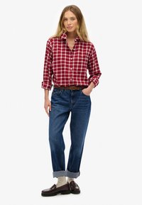 Chemise à carreaux rouge et blanc avec manches retroussées, associée à un jean bleu tapered. Une ceinture en cuir marron et des mocassins bordeaux complètent le look.