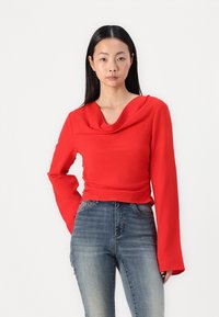 Rød, cowl-hals langermet topp i teksturert stoff. Kombinert med høydejusterte blå denim-jeans. Nøytral bakgrunn.