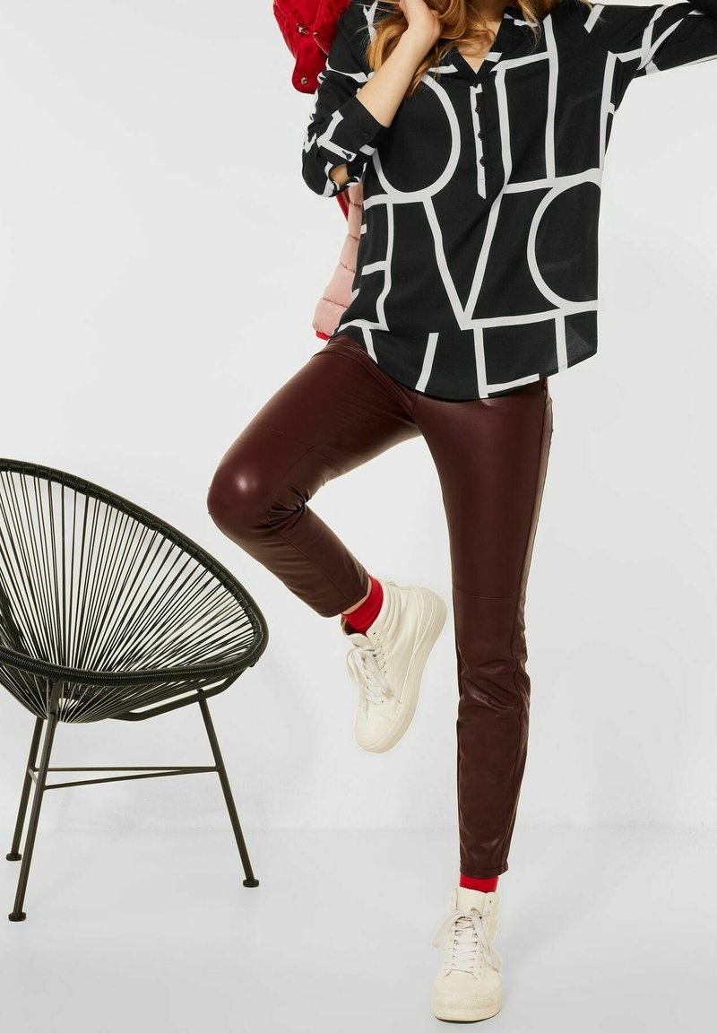 Femme portant un haut à motif noir et blanc et un pantalon en cuir marron, levant une jambe tout en se tenant à côté d'une chaise en fil noir.