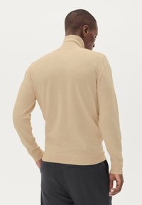 Pulover bej cu guler înalt, realizat dintr-un material tricotat moale, cu mâneci lungi și mansete cu striații, având un design ajustat, purtat cu pantaloni închiși la culoare.