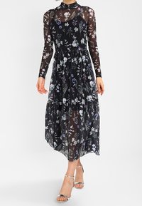 Robe midi noire à motifs floraux avec de longues manches transparentes et un col haut. Caractérisée par une jupe à volants et des motifs floraux mélangés dans des nuances de violet.