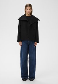 Oversized sort uldmix frakke med bred krave og to store knapper, parret med løstsiddende mørkeblå denimbukser.