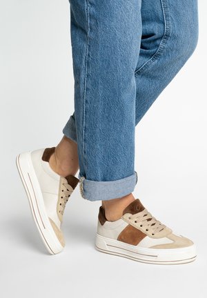 Witte en bruine sneakers gedragen met opgerolde blauwe spijkerbroeken, één voet iets opgetild, op een effen lichte achtergrond.