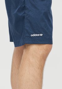 Μπλε σκούρο σορτς Adidas που φοριούνται πάνω από το γόνατο με ορατό λογότυπο της μάρκας στο πλάι σε απλό φόντο.