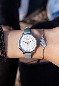 Silverfärgad klocka med en strukturerad tusenskönaface och ljusblå läderrem. Silverarmband med tre tusensköna-liknande detaljer.