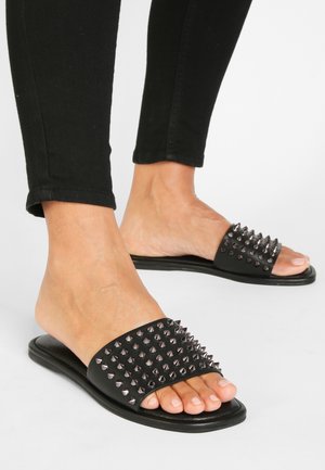 Les sandales à glissière noires présentent une large bride ornée de pointes métalliques argentées. La semelle est plate, avec une texture lisse et un design minimaliste.