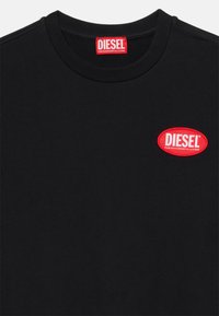 T-shirt in cotone nero con scollo rotondo. Presenta una toppa ovale rossa con il testo "DIESEL" e uno slogan sottostante. Tessuto morbido, design semplice.