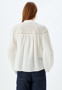 Chemisier blanc léger avec un design en œillets au niveau du corsage, manches bouffantes et dos froncé. Associé à un denim bleu foncé.