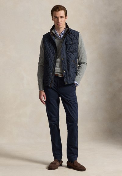 Hombre vistiendo chaleco acolchado azul marino, suéter gris, camisa a rayas, pantalones azul marino y mocasines marrones, de pie sobre un fondo neutro.