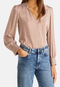 Blus med långa ärmar i ett brunt geometriskt mönster, V-ringning, mjukt tyg, med rynkade axlar, matchad med ljusblå högmidjade jeans.