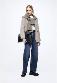 Bēšs rūtainais žakete ar brūnu šalli, balta krekls, zilas plašas bikses un melnas platformas kurpes, turot melnu somu.