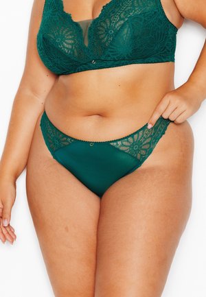Donna che indossa un reggiseno in pizzo verde e mutandine abbinate, tenendo la vita delle mutandine con una mano.