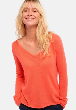 Lächelnde Frau mit langen blonden Haaren, die einen locker sitzenden korallenfarbenen V-Ausschnitt-Pullover und dunkle Hosen trägt, mit den Händen in den Taschen.