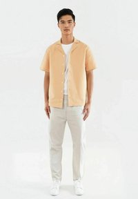 Persikofärgad kortärmad skjorta med en texturerad mönster, knapprad framtill, bärs över en vit t-shirt; matchas med ljusgrå byxor och vita sneakers.