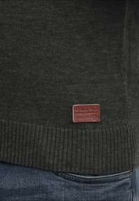 Pull en tricot gris foncé avec un ourlet côtelé, présentant une étiquette en cuir marron embossée avec "BLEND GLOBAL JEANSMAKERS."