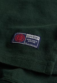 Donkergroene katoenen t-shirt met een blauw en rood stoffen label met de tekst "SUPERDRY VINTAGE ATHLETIC OUTFITTERS." Zachte textuur en gestikte randen.