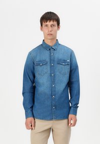 Denim košulja u srednjoj plavoj boji s dugim rukavima, dva džepa na prsima i dugmadi sprijeda. Ima klasičan ovratnik i blagi izblijedjeli izgled.