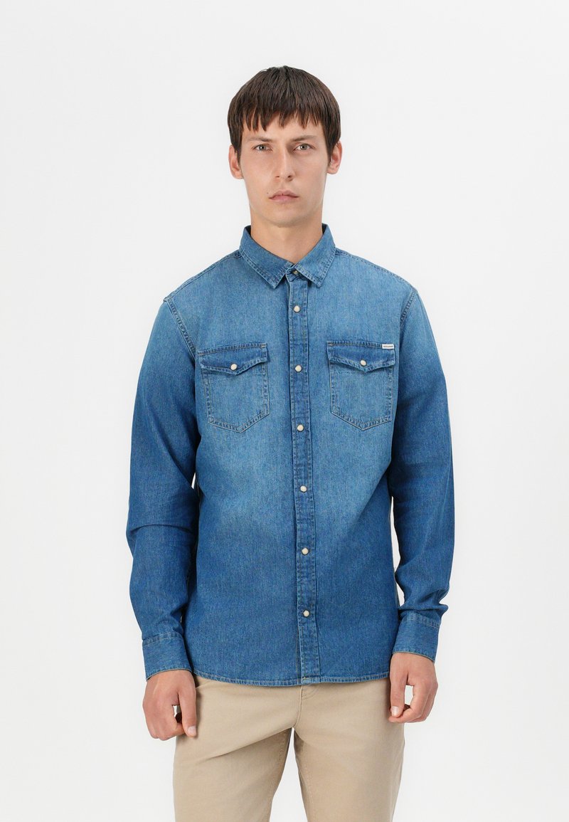 Denim košulja u srednjoj plavoj boji s dugim rukavima, dva džepa na prsima i dugmadi sprijeda. Ima klasičan ovratnik i blagi izblijedjeli izgled.