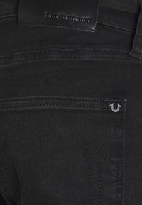 Černé džínsy z denimu s koženou páskovou štítkem s vyraženým nápisem "TRUE RELIGION" a charakteristickým stříbrným logem na zadní kapse.