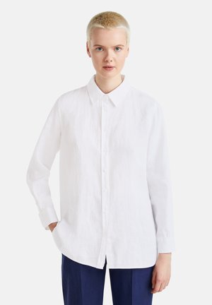 Camisa de lino blanca con cuello puntiagudo, mangas largas y dobladillos sin acabar, combinada con pantalones azul marino. La textura parece ligera y aireada.