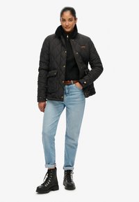 Sorte quilted jakke med høj krave, parret med en sort strikket sweater og lyseblå jeans. Har guldfarvet hardware og lommer. Sorte støvler.