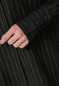 Camicia nera con sottili righe verticali gialle, dotata di polsini con bottoni e tessuto testurizzato. I bottoni sono tondi e neri, aggiungendo un tocco di dettaglio.