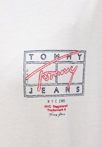 Valkoinen kangas, jossa monivärinen graafinen logo, jossa "TOMMY" ja "JEANS" sinisinä, punaisin aksentein ja pieni teksti "NYC 1985" alapuolella.
