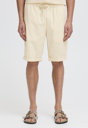 Light yellow cotton shorts with an elastic waistband, drawstring, and rolled cuffs. Worn with brown sandals, featuring double straps.
