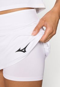 Bela športna obleka, ki vključuje krilo in prilegajoče hlače spodaj. Material je gladek, na hlačah je črn logotip.