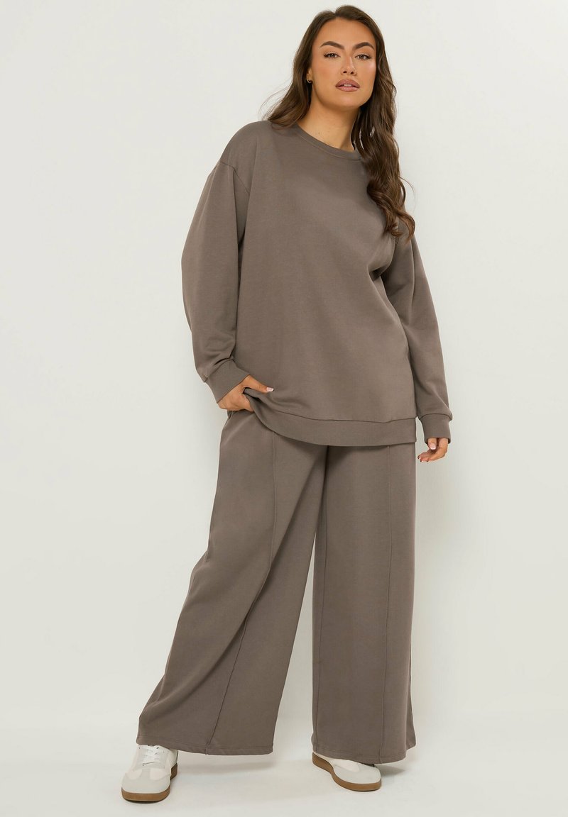 Kvinde iført løs taupe sweatshirt og bukser med vide ben samt hvide sneakers, stående mod en ensfarvet lys baggrund.