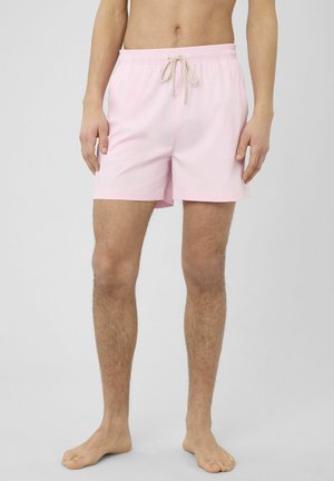 Homme portant un short de bain rose clair avec cordon de serrage, debout pieds nus devant un fond gris uni.