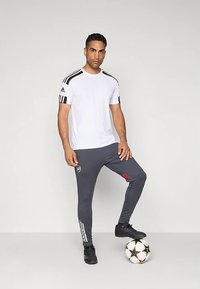 Camiseta deportiva blanca con rayas negras de Adidas, combinada con pantalones grises entallados con detalles rojos y rayas blancas. Botas de fútbol negras.