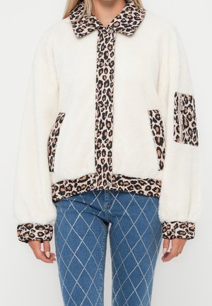 Femme portant une veste polaire blanche avec un bord à imprimé léopard et un jean bleu avec un motif en losange blanc.