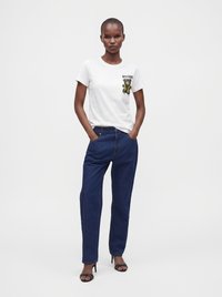 Γυναίκα που φοράει λευκό t-shirt Moschino με στάμπα αρκουδάκι, σκούρα μπλε ψηλόμεσα τζιν και μαύρα πέδιλα με ψηλά τακούνια, τα χέρια στις τσέπες.