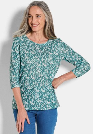 Türkis gemusterte Bluse mit Dreiviertelärmeln, versehen mit einem Diamantdesign in weißen Akzenten. Weicher Stoff mit lässiger Passform. Kombiniert mit blauen Jeans.