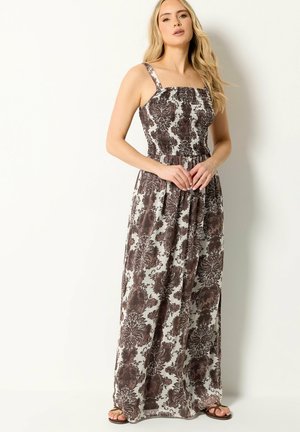 Vrouw draagt een mouwloze, bruine maxi-jurk met bloemenpatroon en een geplooide buste en sandalen, staand tegen een eenvoudige witte achtergrond.