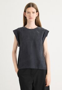 Canotta grigia scura in morbida suede con scollo rotondo e spalle strutturate; abbinata a pantaloni neri, design minimalista.