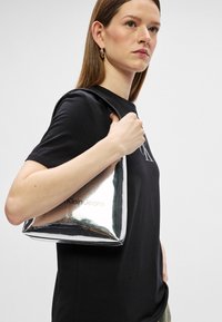 Srebrna metalik torba s sjajnom površinom i crnim detaljima, na kojoj se nalazi natpis "Calvin Klein Jeans." Glatka tekstura i pravokutni oblik.