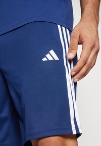 Shorts de sport bleu marine avec trois rayures blanches sur le côté, portant un logo Adidas blanc. Fabriqués en tissu lisse et texturé.