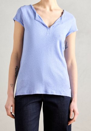 Vrouw die een lichtblauw shirt met korte mouwen en een V-hals draagt en donkere spijkerbroek, met kleine tatoeages op beide onderarmen zichtbaar.