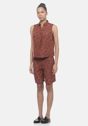 Donna in piedi che indossa un top marrone senza maniche con colletto abbinato e pantaloncini al ginocchio con motivo floreale scuro e mocassini neri.