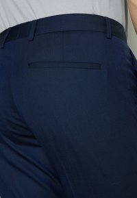 Navy blue -väriset räätälöidyt housut, joissa on sileä pinta, takatasku ja litteä vyötärö. Kangas on hieno kudos ja sillä on hienovarainen kiilto.