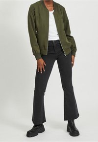 Blouson aviateur vert olive avec fermeture éclair à l'avant, associé à un jean noir ajusté à jambe évasée et des bottines noires à cheville. Tissu lisse et manches retroussées.